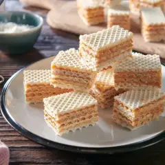 Słodkie wafle przełożone kremową masą do wafli, ułożone na talerzu. Obok słoiczek z masą i miseczka z wiórkami.