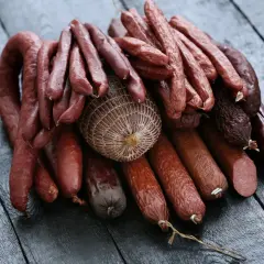 Stos różnych wędlin, w tym grube serdelki, cienkie kiełbaski i wędzonka w siatce, ułożone na ciemnym, drewnianym blacie.