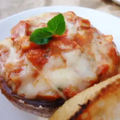 Pyszne pieczarki z grilla z ciągnącą się mozzarellą i sosem pomidorowym, udekorowane listkiem bazylii. Obok kromka chleba.