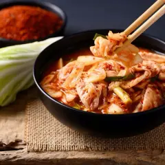 Miseczka pełna pikantnego kimchi, z pałeczkami podnoszącymi kawałek kapusty. Obok liść kapusty i miseczka z przyprawami. To właśnie kimchi!