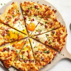 Kawałki pizzy carbonara z jajkiem sadzonym, boczkiem i szczypiorkiem, ułożone na desce.