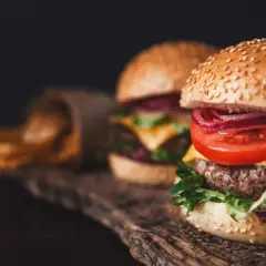 Dwa soczyste smashburgery z serem, pomidorem i czerwoną cebulą, obok kubełka z frytkami.