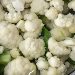 Jak ugotować kalafior? Białe różyczki kalafiora gotują się w garnku z wodą i zielonymi liśćmi.