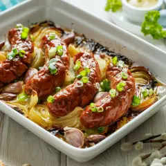 Pieczona biała kiełbasa surowa z cebulą i czosnkiem w naczyniu żaroodpornym, posypana szczypiorkiem.