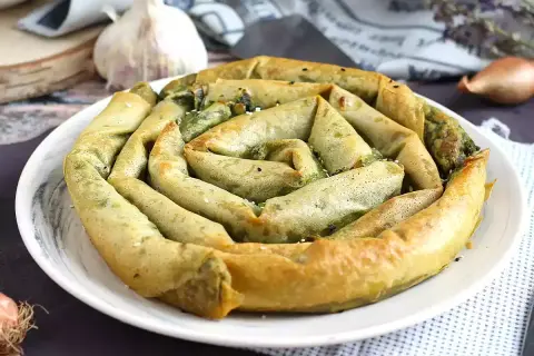 Złocisty borek ze szpinakiem, zwinięty w spiralę, prezentuje się apetycznie na białym talerzu. W tle czosnek i cebulka.