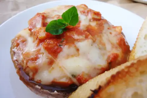 Pyszne pieczarki z grilla z ciągnącą się mozzarellą i sosem pomidorowym, udekorowane listkiem bazylii. Obok kromka chleba.