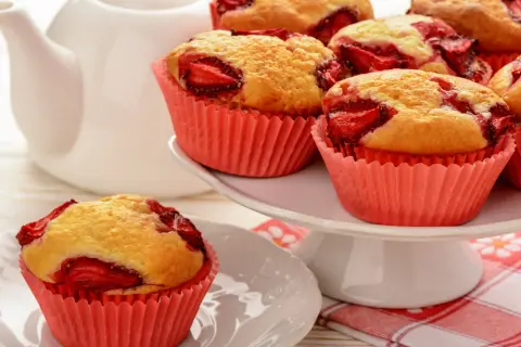 Pyszne muffiny z truskawkami w różowych papilotkach, gotowe do podania z białą czajniczkiem w tle.