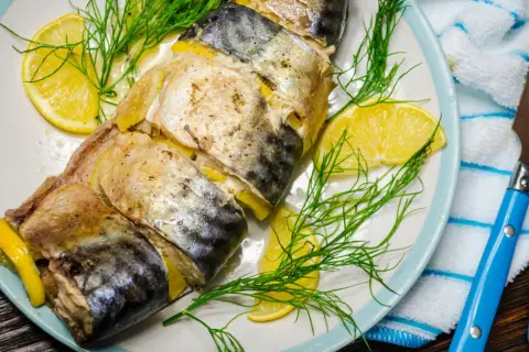 Pieczona makrela z cytryną i koperkiem na talerzu, gotowa do podania z niebieskimi sztućcami.