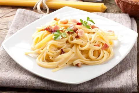 Pyszna carbonara z boczkiem, udekorowana natką pietruszki, na białym talerzu.