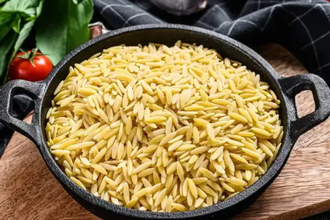 Czarny garnek pełen makaronu w kształcie ryżu, obok pomidory i zielenina.