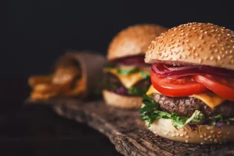 Dwa soczyste smashburgery z serem, pomidorem i czerwoną cebulą, obok kubełka z frytkami.