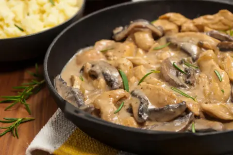 Pyszny kurczak w sosie pieczarkowym, jak u Ani gotuje, w czarnej patelni, z gałązkami rozmarynu. Obok miska z purée.
