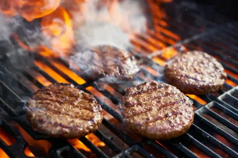 Aromatyczne burgery z grilla skwierczą na rozżarzonych węglach, otoczone dymem i płomieniami.