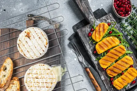 Dwa krążki serów na grilla, jeden lekko roztopiony, obok grzanki. Na desce obok grillowane kawałki dyni z rukolą i granatem.