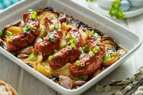 Pieczona biała kiełbasa surowa z cebulą i czosnkiem w naczyniu żaroodpornym, posypana szczypiorkiem.
