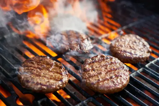 Aromatyczne burgery z grilla skwierczą na rozżarzonych węglach, otoczone dymem i płomieniami.