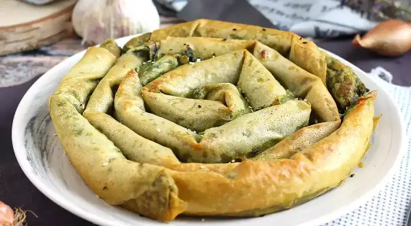 Złocisty borek ze szpinakiem, zwinięty w spiralę, prezentuje się apetycznie na białym talerzu. W tle czosnek i cebulka.