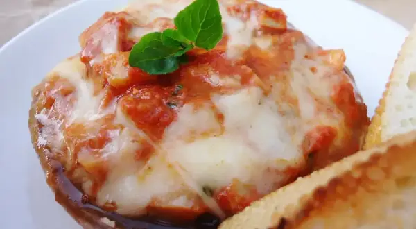 Pyszne pieczarki z grilla z ciągnącą się mozzarellą i sosem pomidorowym, udekorowane listkiem bazylii. Obok kromka chleba.