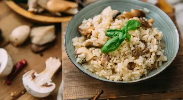 Aksamitny ryż do risotto z grzybami, udekorowany listkiem bazylii. Na drewnianej desce widać świeże grzyby i papryczkę chili.
