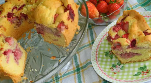 Puszysta babka z truskawkami, pachnąca latem, na szklanej paterze. Obok miseczka świeżych owoców i kawałek ciasta na talerzyku.