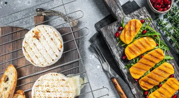 Dwa krążki serów na grilla, jeden lekko roztopiony, obok grzanki. Na desce obok grillowane kawałki dyni z rukolą i granatem.