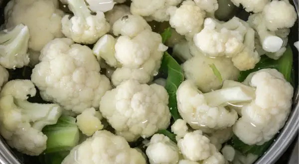 Jak ugotować kalafior? Białe różyczki kalafiora gotują się w garnku z wodą i zielonymi liśćmi.