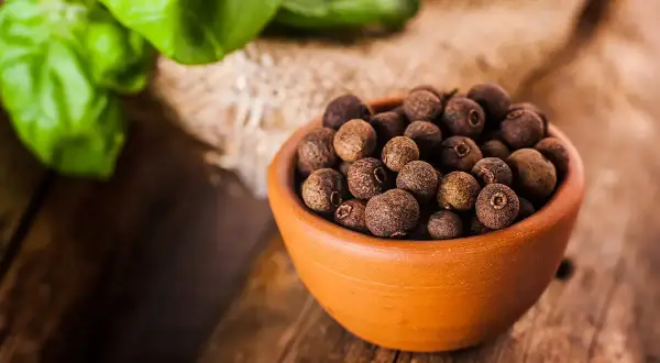 Garnek pełen aromatycznego ziela angielskiego na drewnianym blacie, obok zielone liście.