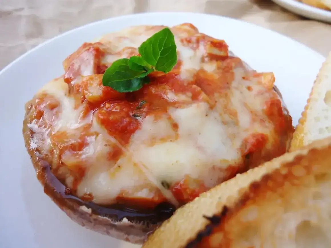 Pyszne pieczarki z grilla z ciągnącą się mozzarellą i sosem pomidorowym, udekorowane listkiem bazylii. Obok kromka chleba.