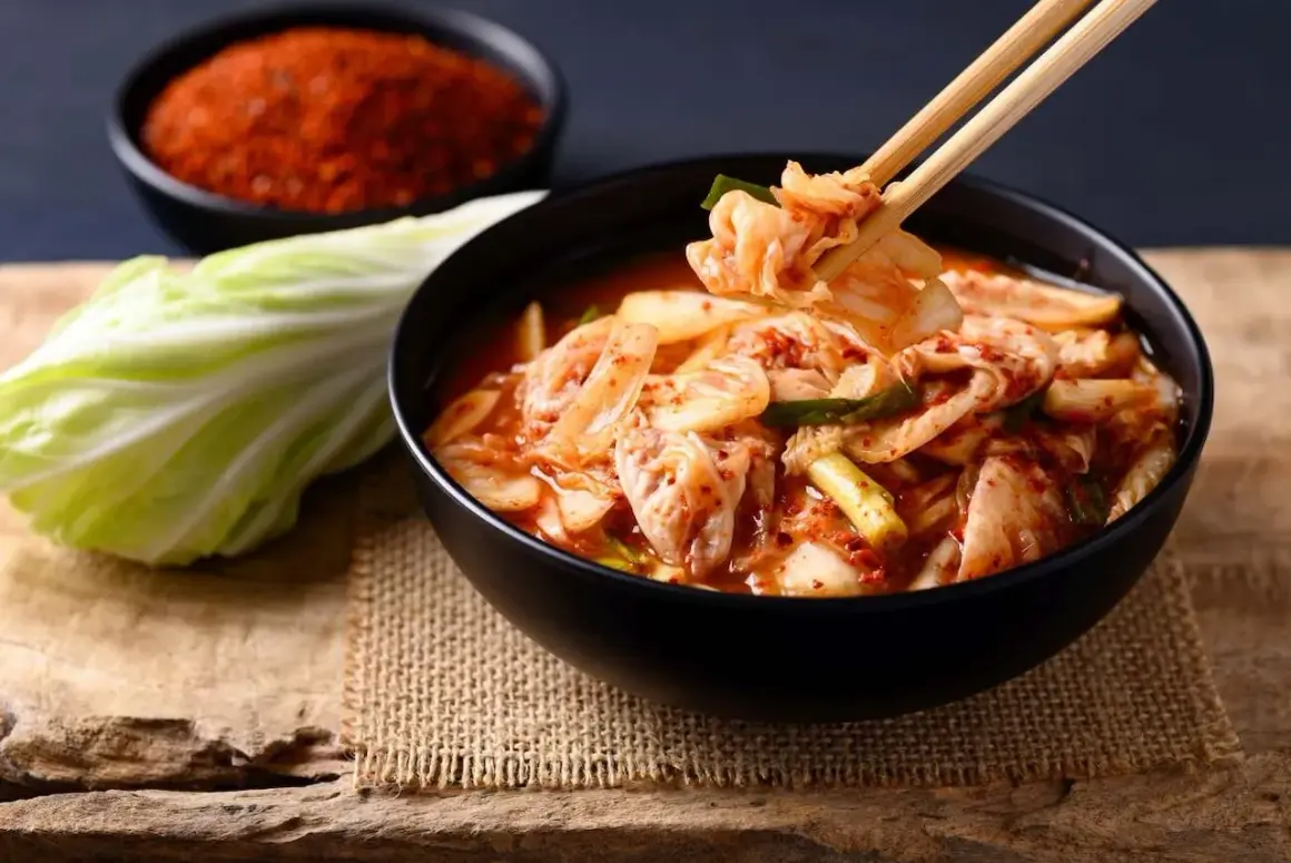 Miseczka pełna pikantnego kimchi, z pałeczkami podnoszącymi kawałek kapusty. Obok liść kapusty i miseczka z przyprawami. To właśnie kimchi!