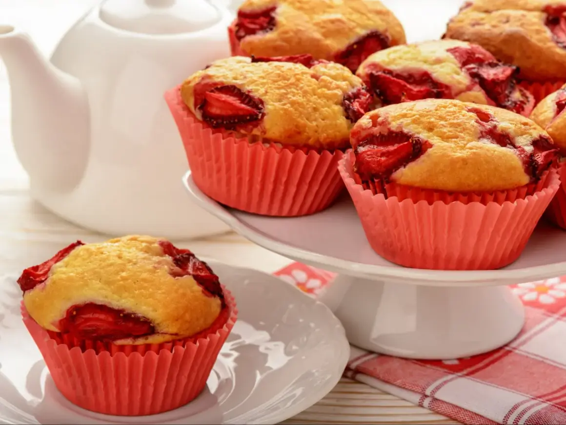 Pyszne muffiny z truskawkami w różowych papilotkach, gotowe do podania z białą czajniczkiem w tle.