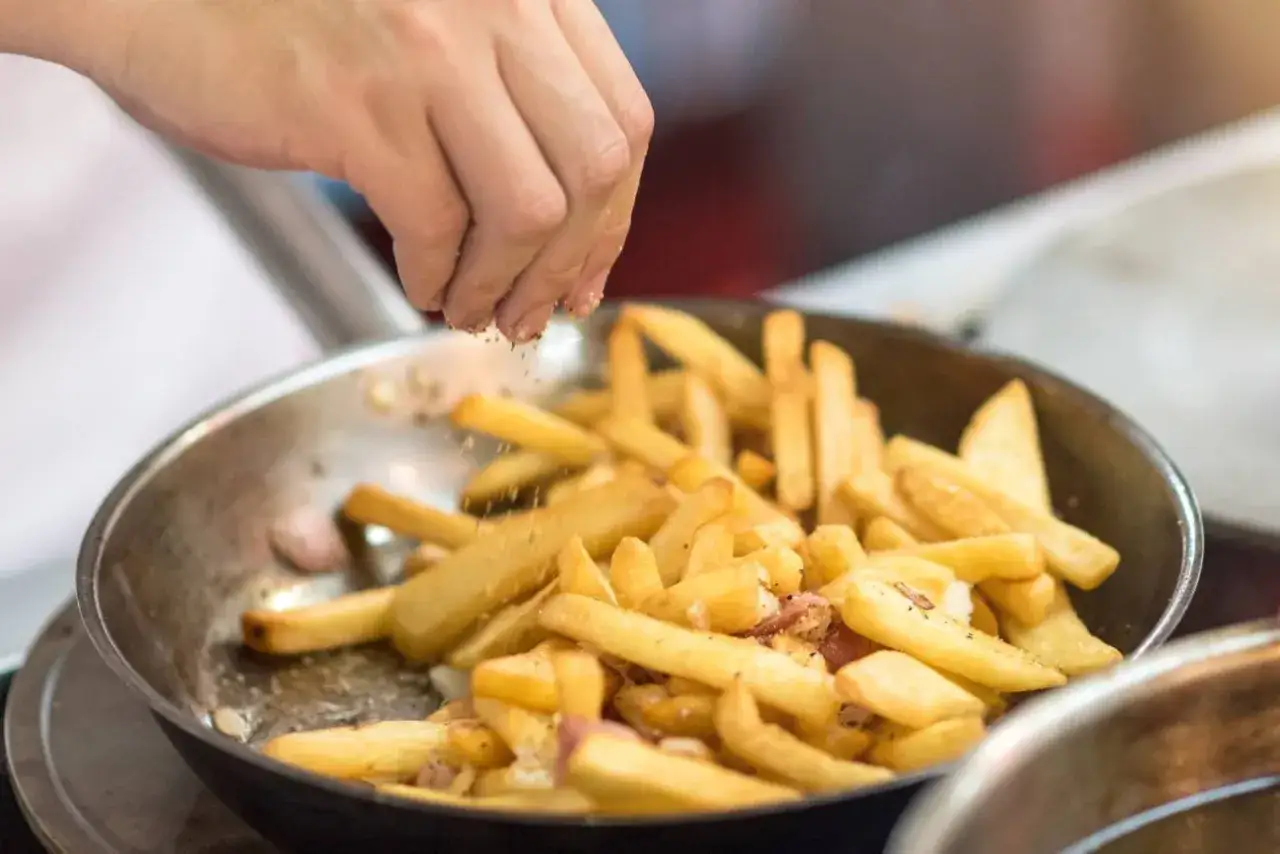 Ręka posypuje przyprawami złociste frytki z airfryer w metalowej misce.