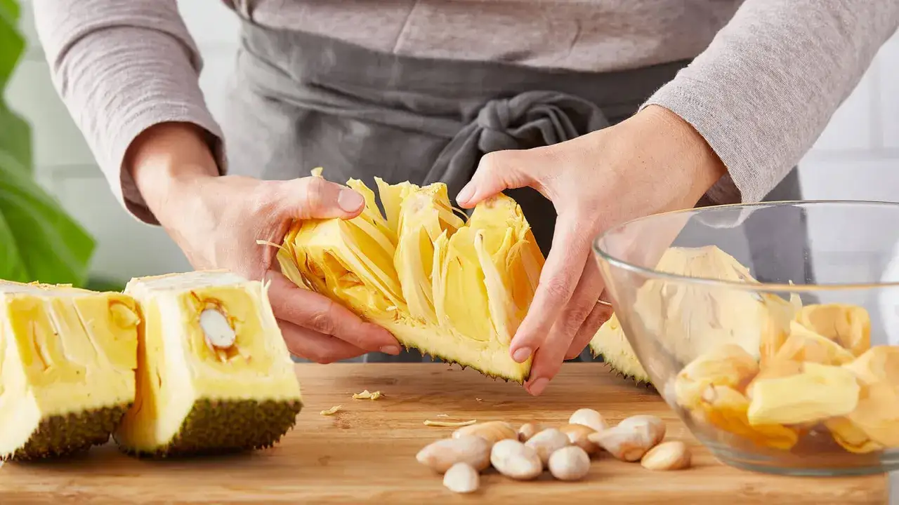Ręce rozdzielają żółte, włókniste części owocu jackfruit. Na desce leżą nasiona i kawałki owocu. Jackfruit co to? To egzotyczny przysmak!
