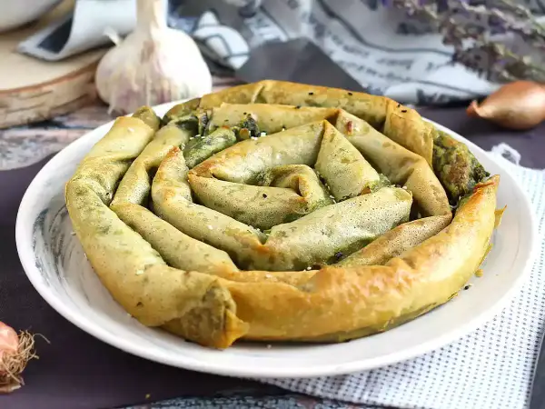 Złocisty borek ze szpinakiem, zwinięty w spiralę, prezentuje się apetycznie na białym talerzu. W tle czosnek i cebulka.