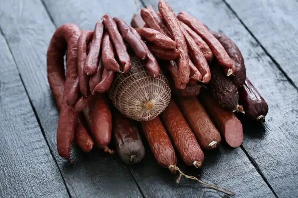 Stos różnych wędlin, w tym grube serdelki, cienkie kiełbaski i wędzonka w siatce, ułożone na ciemnym, drewnianym blacie.