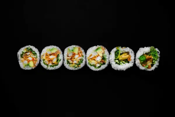 Sześć kawałków futomaki, czyli dużych rolek sushi, z awokado, warzywami i chrupiącym nadzieniem, ułożonych na czarnym tle.