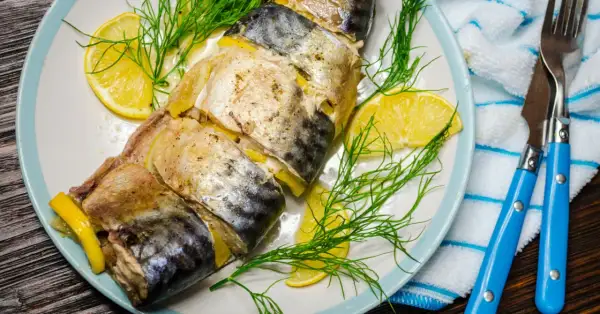 Pieczona makrela z cytryną i koperkiem na talerzu, gotowa do podania z niebieskimi sztućcami.