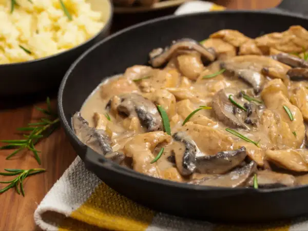 Pyszny kurczak w sosie pieczarkowym, jak u Ani gotuje, w czarnej patelni, z gałązkami rozmarynu. Obok miska z purée.