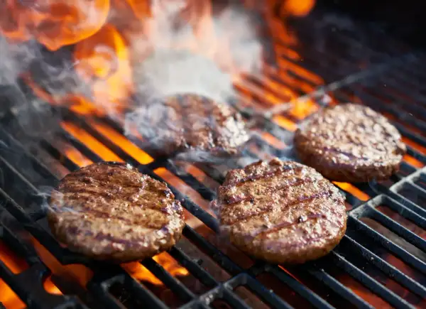 Aromatyczne burgery z grilla skwierczą na rozżarzonych węglach, otoczone dymem i płomieniami.